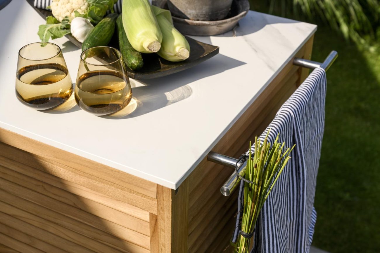 Die Gartenküche Figalia überzeugt mit ihrem modernen Design. Gefertigt wurde sie aus Terrazzo, welches einen weißen Farbton besitzt. Das Gestell ist aus Teakholz und hat eine natürliche Farbe. Das Element verfügt über ein Waschbecken, welches an einen Gar